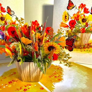 Puede incluir: Una tarjeta de felicitación emergente 3D con un vibrante arreglo floral en una maceta con aspecto de madera. El ramo incluye girasoles, amapolas y lirios en tonos amarillos, naranjas y rojos. Mariposas con alas naranjas y negras revolotean.