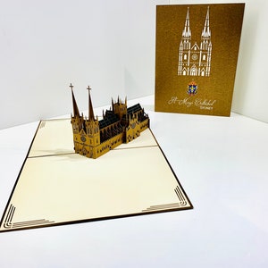 Puede incluir: Una tarjeta pop-up 3D marrón y dorada que representa la Catedral de Santa María en Sídney, Australia. La tarjeta está abierta y la catedral está de pie. La parte delantera de la tarjeta tiene un fondo marrón con un contorno blanco de la catedral y el texto "St. Mary's Cathedral SYDNEY".