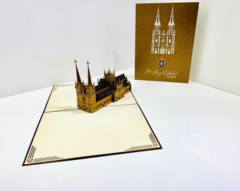 Puede incluir: Una tarjeta pop-up 3D marrón y dorada que representa la Catedral de Santa María en Sídney, Australia. La tarjeta está abierta y la catedral está de pie. La parte delantera de la tarjeta tiene un fondo marrón con un contorno blanco de la catedral y el texto "St. Mary's Cathedral SYDNEY".