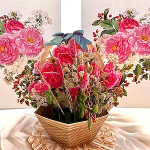 May include: A pop-up floral arrangement featuring pink roses, white flowers, and red berries in a woven basket. A blue ribbon adorns the top. The background includes two floral illustrations.