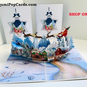 May include: A pop-up Christmas card featuring a winter village scene with Santa Claus in his sleigh, pulled by reindeer. The card includes a star-shaped design with a winter village scene. The text "SHOP ONLINE" is visible.