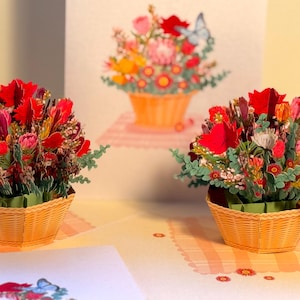 May include: Three-dimensional paper art featuring vibrant floral arrangements in woven baskets. The flowers are a mix of red, pink, and yellow, with green foliage. A butterfly is visible on one of the cards. The background includes a similar floral design.
