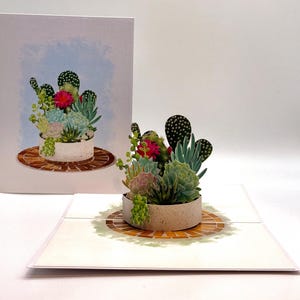 May include: A 3D pop-up greeting card featuring a detailed arrangement of succulents in a round, light-colored pot. The card is open, revealing the pop-up design, with a matching illustration on the front. The succulents include green, pink, and blue varieties.