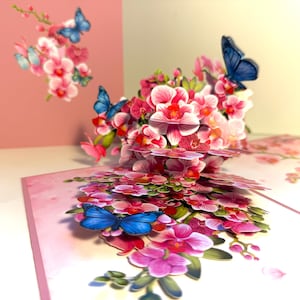 Puede incluir: Una tarjeta de felicitación emergente con orquídeas rosas y blancas y mariposas azules. La tarjeta está abierta, revelando un arreglo floral tridimensional. Una segunda tarjeta con un diseño similar está en el fondo.