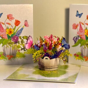 May include: A 3D pop-up greeting card with a vibrant floral display in a wooden basket, accompanied by butterflies. Two additional cards with similar floral designs and butterflies are also present. The flowers include pink, orange, yellow, and purple blooms.