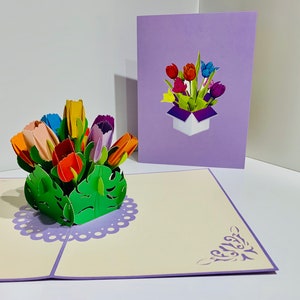Puede incluir: Una tarjeta pop-up 3D con un ramo de tulipanes coloridos en una caja morada. La tarjeta está abierta y los tulipanes están saliendo de la tarjeta. La tarjeta está sobre una superficie blanca.