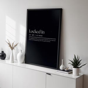 Puede incluir: Un póster enmarcado en negro con el texto "Locked In" y su definición. El póster se exhibe en un mueble blanco, junto a jarrones blancos y negros, una pequeña planta y una lámpara decorativa. La habitación tiene un diseño minimalista.