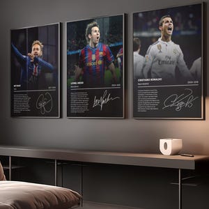 Puede incluir: Tres carteles enmarcados con las estrellas de fútbol Neymar, Lionel Messi y Cristiano Ronaldo. Cada cartel tiene una foto del jugador, su nombre, equipo, años jugados y una firma. Los carteles están colgados en una pared gris oscura.