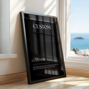 May include: Black framed art print with the word "CUSTOM" at the top and "CUSTOM 2:3" below. The quote "But you Lord, are the shield around me, my glory, the one who lifts my head high" is at the bottom. The print is leaning against a wall.