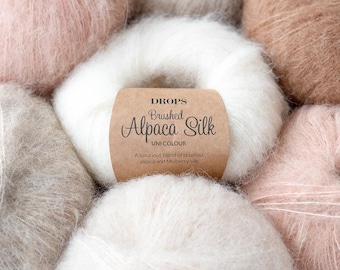 May include: A collection of soft, fuzzy yarn balls in various neutral and pastel shades. A central white ball features a tan paper label with the text DROPS Brushed Alpaca Silk UNI COLOUR, A luxurious blend of brushed alpaca and Mulberry silk.