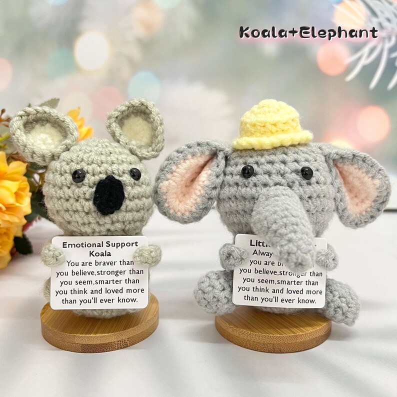 May include: Two crocheted stuffed animals, a koala and an elephant, are displayed on wooden bases. The koala is holding a card that reads "Emotional Support Koala" and the elephant is holding a card that reads "Little Always You are..."