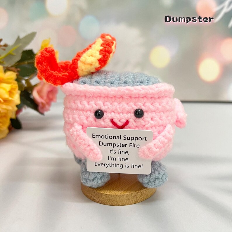 May include: A pink and gray crocheted plush toy shaped like a dumpster fire with a smiling face. The toy is holding a sign that reads "Emotional Support Dumpster Fire It's fine, I'm fine. Everything is fine!"