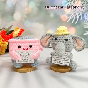 May include: Two crocheted plush toys, a pink dumpster fire and a gray elephant, each holding a card with a message. The dumpster fire card reads "Emotional Support Dumpster Fire It's fine, I'm fine. Everything is fine!" The elephant card reads "Remember you believe stronger than you seem, smarter than you think and loved more than you'll ever know."