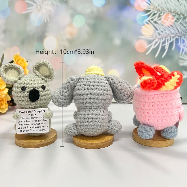 May include: Three crocheted stuffed animals on wooden bases. The animals are a gray koala, a gray elephant, and a pink and orange fox. The koala is holding a card that reads "Emotional Support Koala. You are braver than you believe, stronger than you seem, smarter than you think and loved more than you'll ever know."