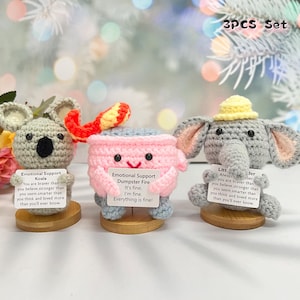 May include: Three crocheted stuffed animals on wooden bases. The animals are a koala, a dumpster fire, and an elephant. Each animal has a small sign in front of it with an encouraging message. The signs read "Emotional Support Koala", "Emotional Support Dumpster Fire", and "Litt Always Jer ber".
