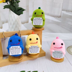 May include: Four crocheted dinosaur toys, each a different color: green, blue, yellow, and pink. Each dinosaur has a small tag attached with the text "Emotional Support Dinosaur. Be brave, be curious, stand tall and dream big. Let the world hear you roar!"