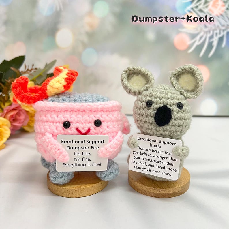 May include: Two crocheted plushies, one shaped like a dumpster fire and the other like a koala, each holding a sign with encouraging messages. The dumpster fire plush is pink with a gray top and a red flame on top. The koala plush is gray with white ears and a black nose. The signs read "Emotional Support Dumpster Fire It's fine, I'm fine. Everything is fine!" and "Emotional Support Koala You are braver than you believe, stronger than you seem, smarter than you think and loved more than you'll ever know."