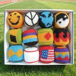 Puede incluir: Una caja de doce sacos de hacky tejidos de colores. Cada saco presenta un diseño único, incluyendo una carita sonriente, un signo de la paz, un símbolo del yin-yang y la bandera estadounidense. Los sacos están en una caja blanca sobre un campo de hierba verde.
