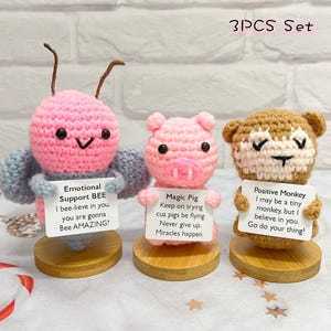 Könnte beinhalten: Set aus drei gehäkelten Tierfiguren mit motivierenden Botschaften. Eine rosa Biene mit grauen Flügeln hält ein Schild mit der Aufschrift "Emotional Support BEE I bee-lieve in you, you are gonna Bee AMAZING!" Ein rosa Schwein hält ein Schild mit der Aufschrift "Magic Pig Keep on trying cuz pigs be flying. Never give up. Miracles happen." Ein brauner Affe hält ein Schild mit der Aufschrift "Positive Monkey I may be a tiny monkey, but I believe in you. Go do your thing!"