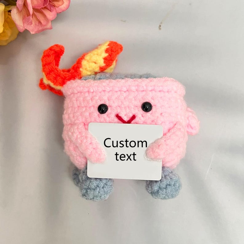 May include: A pink crocheted character with a yellow and orange flame on top, holding a white card with the text "Custom text".