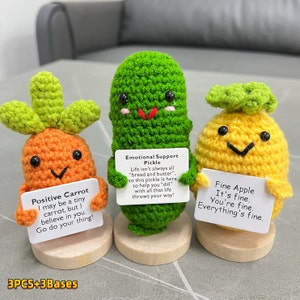 May include: Three crocheted plushies, a carrot, a pickle, and an apple, each holding a sign with a positive message. The carrot says "Positive Carrot I may be a tiny carrot, but I believe in you. Go do your thing!" The pickle says "Emotional Support Pickle Life isn't always all "bread and butter", so this pickle is here to help you "dill" with all that life throws your way!" The apple says "Fine Apple It's fine. You're fine. Everything's fine."