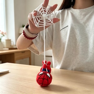 May include: A white crocheted spider web accessory attached to a long white yarn string, which suspends a small, round, red and blue crocheted character with white eye patches and black web-like detailing on its surface.