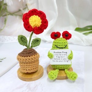 May include: A crocheted green frog holding a sign that reads "Positive Frog I may be a tiny frog, but I believe in you. Go do your thing!" The frog is sitting on a small wooden base. There is also a crocheted red flower in a brown pot on a wooden base.