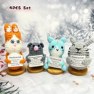 May include: Set of four crocheted cat plushies, each holding a sign with a positive message. The plushies are orange, gray, blue, and gray. The signs read "Positive Cat", "Emotional Support Mouse", "Positive Cat", and "Positive Cat".