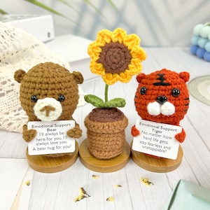 May include: Crocheted stuffed animals with motivational messages. A brown bear holds a sign that says "Emotional Support Bear I'll always be here for you. I'll always love you. A bear hug for you!" A crocheted sunflower plant is in a brown pot. A tiger holds a sign that says "Emotional Support Tiger No matter how hard life gets, stay fearless and unstoppable!"