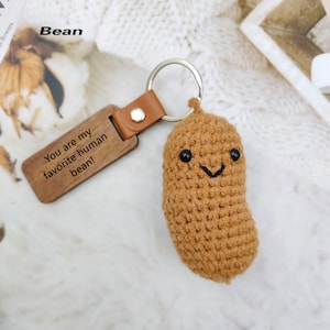 May include: A brown crocheted bean-shaped keychain with a smiling face and a wooden tag. The tag reads "You are my favorite human bean!" The keychain has a leather strap and a silver keyring.