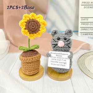 May include: Two crocheted amigurumi figures: a gray mouse holding a sign that reads "Emotional Support Mouse All our dreams can come true if we have the courage to pursue them." and a sunflower in a brown pot. Both figures are on wooden bases.