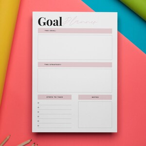 Könnte beinhalten: Ein weißer Zielplaner mit den Worten "Goal Planner" oben. Der Planer hat Abschnitte für "The Goal", "The Strategy", "Steps to Take" und "Notes". Der Planer liegt auf einer rosa Oberfläche mit gelb und blau gerollten Papier auf beiden Seiten.