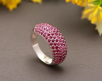 Puede incluir: Un anillo de plata con una banda ancha engastada con pequeñas piedras preciosas de color rojo oscuro. El anillo está sobre una superficie gris.