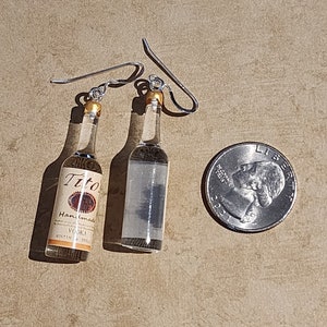 May include: A pair of silver dangle earrings featuring miniature glass bottles. One bottle is clear and the other has a label that reads "Tito's Handmade Vodka".