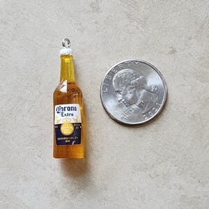 May include: A miniature glass bottle charm with a gold cap and a label that reads "Corona Extra". The bottle is filled with a yellow liquid and is about the size of a US quarter.