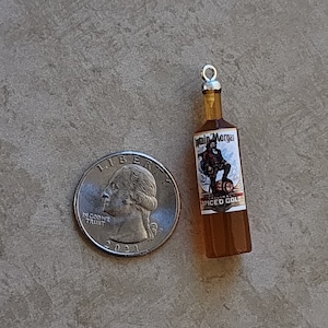 May include: A miniature glass bottle charm with a silver cap and a label that reads "Captain Morgan Original Spiced Gold". The bottle is filled with a dark amber liquid and features a pirate illustration on the label.