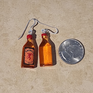 May include: A pair of silver earrings shaped like miniature Fireball whiskey bottles. The bottles are clear with a red cap and a label that says "Fireball" with a red devil on it.