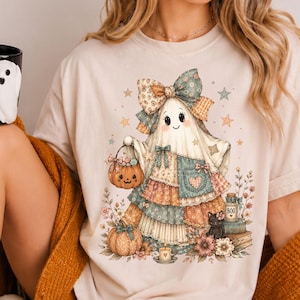 May include: A beige t-shirt featuring a graphic of a ghost wearing a patchwork dress and a large bow, holding a pumpkin bucket. The ghost is surrounded by flowers, pumpkins, and a small black cat. The shirt is worn with cream-colored socks.
