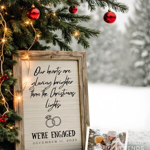 Pode incluir: Uma placa de madeira com o texto "Our hearts are glowing brighter than the Christmas lights" e "We're Engaged December 11, 2025" é exibida em frente a uma árvore de Natal com enfeites vermelhos e luzes de corda. Uma pequena foto está no canto inferior direito.