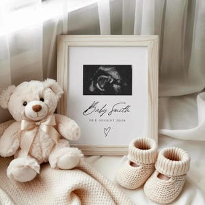 Pode incluir: Uma imagem de ultrassom emoldurada com o texto "Baby Smith DUE AUGUST 2026" e um símbolo de coração. Ao lado da moldura, um urso de pelúcia creme com um laço e um par de botinhas de bebê de malha.