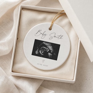 May include: A round white ceramic ornament featuring a black and white ultrasound image in the centre. The text Baby Smith is printed above the image, and a small heart icon with the text JUNE 2027 is printed below. It hangs by a gold cord.
