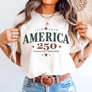 Pode incluir: Camiseta creme com a palavra "AMERICA" em verde escuro e o número "250" em vermelho. A camisa também apresenta os anos "1776 2026" e "YEARS OF FREEDOM" em um estilo vintage. Um cinto marrom com fivela decorativa é visível.