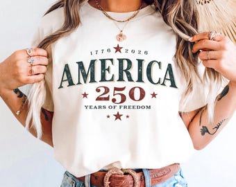 Op de afbeelding: Crèmekleurig T-shirt met het woord "AMERICA" in donkergroen en het nummer "250" in rood, ter viering van 250 jaar vrijheid. Het shirt toont de jaartallen "1776" en "2026" boven het woord "AMERICA".