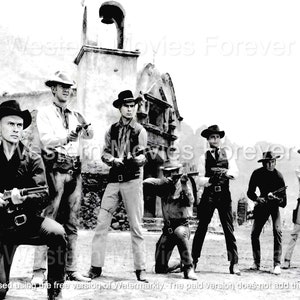 以下が含まれることがあります： 西部劇のセットにいる7人のカウボーイの白黒写真。彼らは皆帽子をかぶり、銃を持っている。カウボーイたちは鐘楼のある建物の前に立っている。