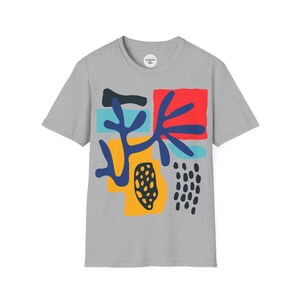 May include: A grey t-shirt with a vibrant abstract design. The design incorporates red, blue, yellow, and black shapes. The t-shirt has a crew neck and short sleeves. A small circular tag is visible near the neckline.