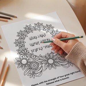 Könnte beinhalten: Eine Ausmalseite mit einem floralen Kranzdesign und hebräischen Text wird mit einem grünen Bleistift ausgemalt. Der Text lautet: "Bitte, mit der Stärke Deiner rechten Hand, gib frei, was gebunden ist." Buntstifte liegen verstreut herum.
