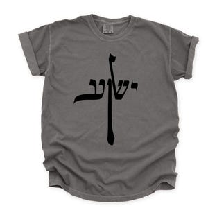 May include: Gray t-shirt with a black Hebrew script design. The shirt has a crew neck and short sleeves with rolled cuffs. The graphic is centered on the chest and appears to be a stylized cross shape.