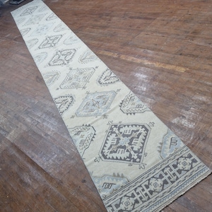 May include: A long, rectangular rug with a cream background and geometric patterns in grey and blue. The rug has a border with a repeating design. Suitable for a hallway or entryway, it measures approximately 300 cm in length.