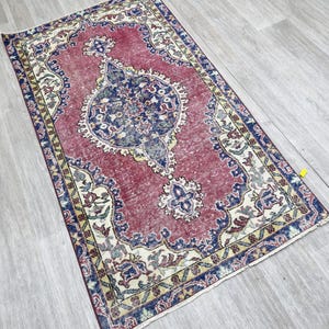 Puede incluir: Una alfombra rectangular con un aspecto vintage y desgastado. La alfombra presenta un diseño central de medallón en azul marino y crema, sobre un fondo rojo descolorido. El borde es una mezcla de azul marino, crema y amarillo, con patrones florales y geométricos.