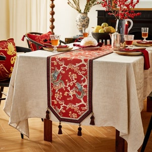 Peut inclure: Un chemin de table rouge et or avec un motif de dragon et de fleurs. Le chemin de table a des glands à chaque extrémité et est sur une nappe beige.